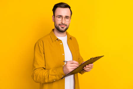 Photo of smart confident young gentleman dressed casual outfit spectacles writing clipboard isolated yellow color backgroundの写真素材