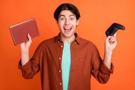 Portrait of young handsome unhappy male choose between joystick book try study isolated on orange...