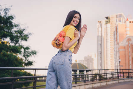 Rear photo of charming positive girl hold fruits net arm palm waving hi look camera pastime outside