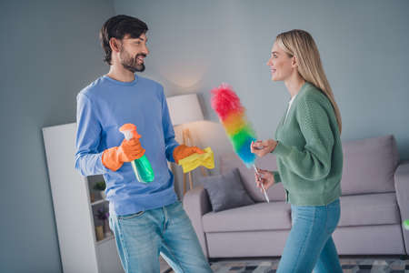 Photo of positive cheerful spouses guy lady clean apartment room hold detergent fluffy duster rag...