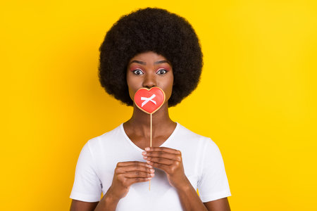 Photo of young funky afro american woman cover mouth candy heart stick isolated on yellow color backgroundの写真素材