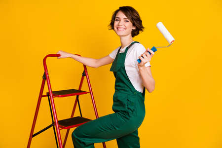 Photo of happy positive smiling girl painter climbing ladder hold roller renovation isolated on yellow color backgroundの写真素材