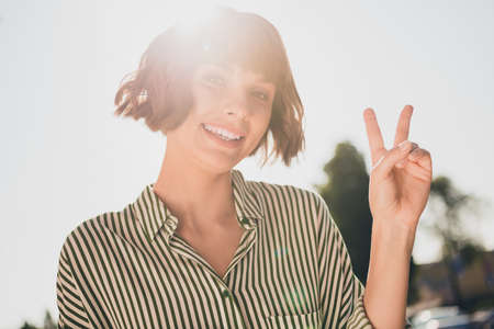 Photo of positive glad friendly cute lady toothy smile show v-sign wear striped shirt urban city outsideの写真素材