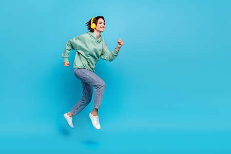 Full length profile photo of cute millennial brunette lady run wear headphones pullover jeans footwear isolated on blue backgroundの写真素材