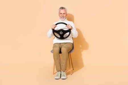 Portrait of attractive cheerful man holding steering wheel sitting in chair learning isolated on...