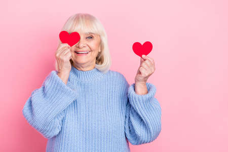 Portrait of attractive cheerful grey-haired woman holding heart cards closing eye isolated over pink...