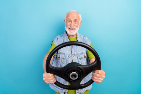 Photo of cute excited retired man wear vintage jeans waistcoat holding steering wheel riding auto smiling isolated blue color backgroundの写真素材