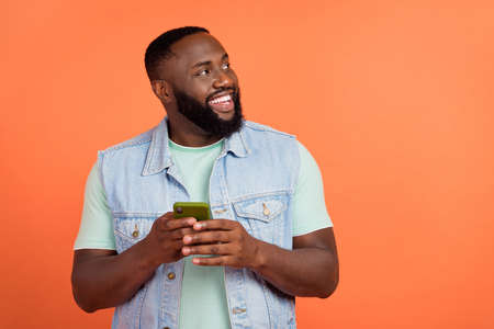 Photo of dreamy guy dressed jeans waistcoat typing gadget looking empty space isolated orange color backgroundの写真素材