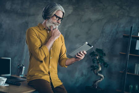 Photo of smart clever mature guy dressed yellow shirt spectacles arm chin reading modern gadget indoors workshop workplaceの写真素材