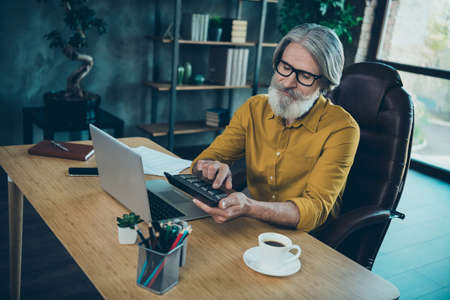 Photo of busy senior man wear yellow shirt glasses working device counting calculator indoors workplace workstationの写真素材