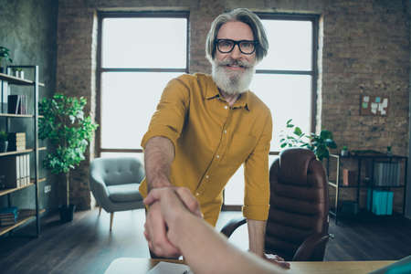 Photo of positive mature guy dressed yellow shirt spectacles handshaking indoors workshop workplaceの写真素材
