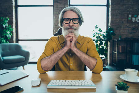 Photo of thoughtful mature guy dressed yellow shirt spectacles looking modern gadget indoors workshop workstationの写真素材
