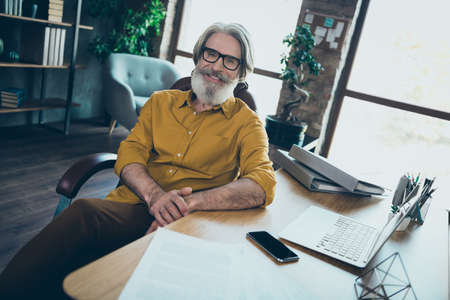 Photo of positive mature guy dressed yellow shirt spectacles working modern gadget indoors workshop workstationの写真素材