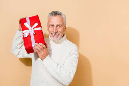 Portrait of attractive cheerful grey-haired man holding gift new year December copy space isolated over beige pastel color backgroundの写真素材