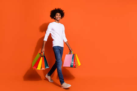 Photo of attractive pretty guy dressed white shirt carrying shoppers walking empty space isolated orange color backgroundの写真素材