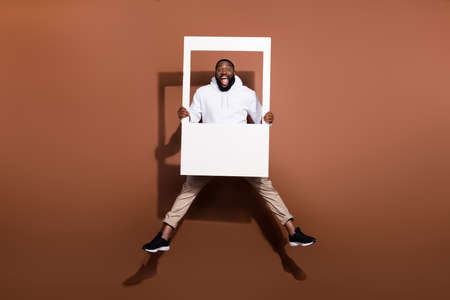 Photo of excited funky guy wear white hoodie jumping high holding paper frame isolated brown color backgroundの写真素材