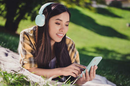 Photo of sweet cute young woman dressed plaid shirt lying blanket green grass listening music modern...