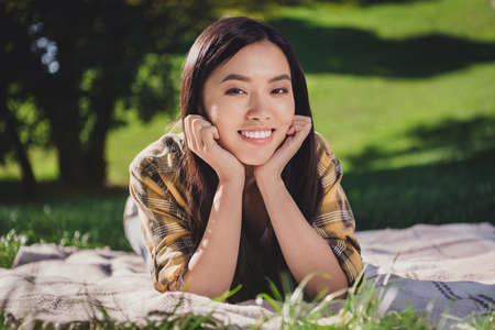 Photo of shiny pretty young lady wear checkered shirt smiling lying green grass blanket outdoors...