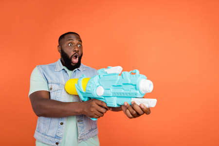 Photo of shocked guy dressed jeans waistcoat shooting water gun empty space isolated orange color...