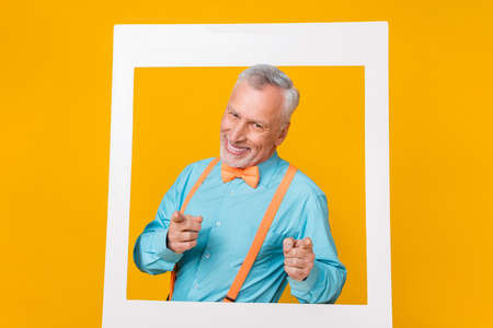 Photo of funky cute retired man wear turquoise shirt suspenders tacking portrait pointing you isolated yellow color backgroundの写真素材