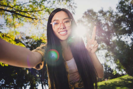 Photo of sweet shiny young lady wear checkered shirt glasses smiling showing v-sign tacking selfie outdoors urban city parkの写真素材