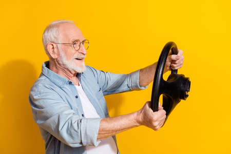 Profile side view portrait of attractive cheerful grey-haired man hold steering wheel having fun isolated on vivid yellow color backgroundの写真素材