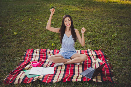 Portrait of attractive cheerful lucky girl sitting on cover using laptop rejoicing having fun on green grass outdoorsの写真素材