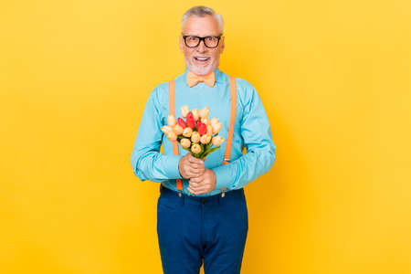 Portrait of attractive cheerful grey-haired man holding flowers congratulate festal occasion isolated over bright yellow color backgroundの写真素材