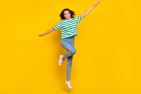 Full body photo of young cheerful girl have fun jump up fly active hands wings isolated over yellow...