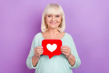 Photo of confident cheerful positive old lady hold icon heart wear teal shirt isolated violet color backgroundの写真素材
