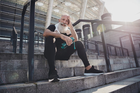 Portrait of attractive healthy cheerful retired grey-haired man working out drinking water sitting on stone stairs active life outdoorsの写真素材