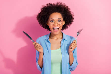 Photo of funny hungry lady hold fork knife prepare eat breakfast open mouth wear denim shirt...
