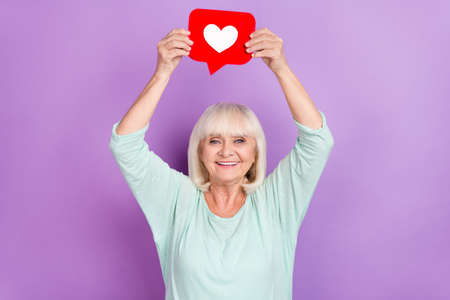 Photo of cheerful funny influencer user old lady hold icon heart raise hands wear teal shirt isolated violet color backgroundの写真素材