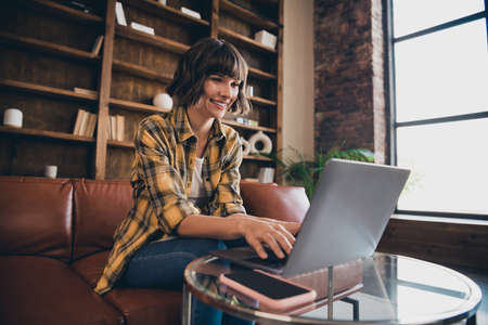 Photo of charming shiny young woman dressed plaid shirt sitting sofa chatting modern gadget indoors workstationの写真素材