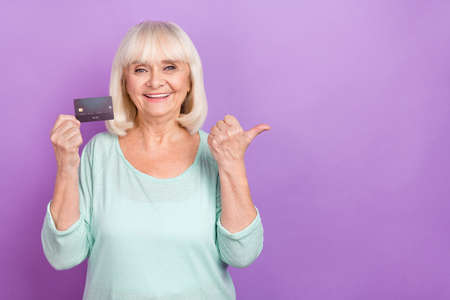 Photo of grandma hold debit card direct finger empty space loan option wear teal shirt isolated violet color backgroundの写真素材
