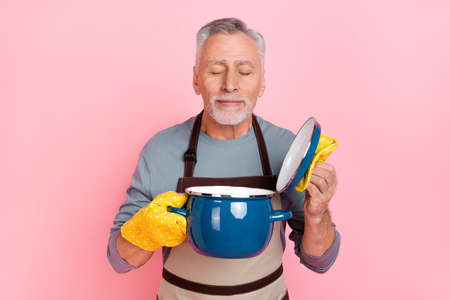 Photo of dreamy funny man pensioner dressed grey shirt apron open saucepan enjoying aroma isolated pink color backgroundの写真素材