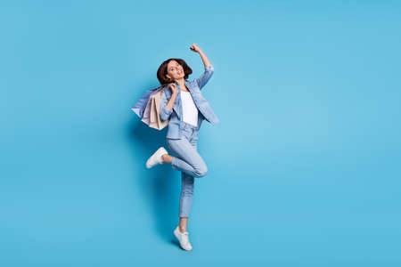 Photo of excited lucky woman wear jeans shirt rising fist bargains jumping high isolated blue color...