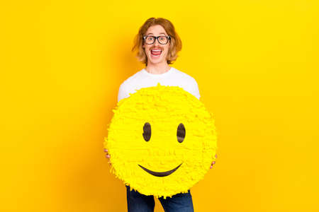 Photo of handsome positive funny male hold big paper pinata smiling emoji isolated on yellow color backgroundの写真素材