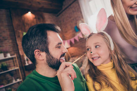 Portrait of positive friendly parents daughter hold tassel paint nose have fun wear rabbit headband...