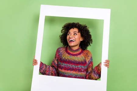 Photo of funky young curly hairstyle lady hold frame look ad wear red sweater isolated on green...