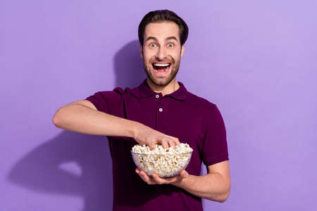 Photo of hooray young brunet guy eat pop corn wear violet polo isolated on purple color backgroundの写真素材
