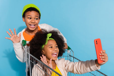 Portrait of small little trendy friends friendship riding cart taking selfie recording video having fun isolated over bright blue color backgroundの写真素材