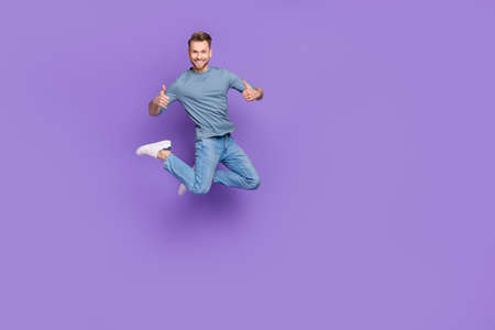 Photo of pretty confident guy wear grey shirt jumping high thumbs up empty space isolated violet...