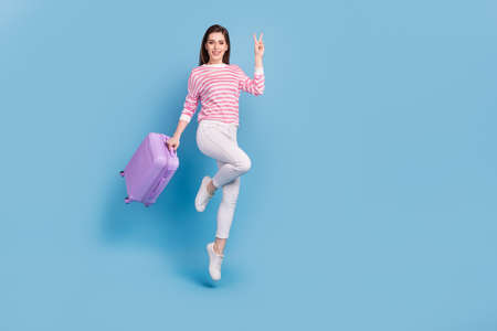 Full size portrait of attractive cheerful lady hold suitcase hand demonstrate v-sign isolated on blue color backgroundの写真素材