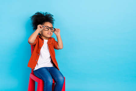 Photo of cute little interested girl sit chair wear eyeglasses thinking dreaming isolated on blue...