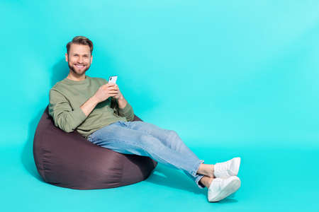 Photo of confident smm blogger man hold telephone sit armchair wear pullover isolated blue color...