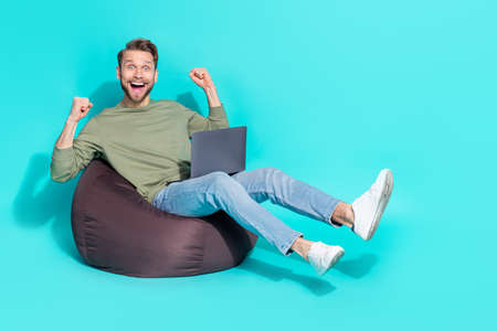 Photo of amazed funny crazy man hold netbook sit armchair rejoice win wear pullover isolated blue color backgroundの写真素材