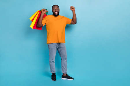 Photo of lucky funky guy wear orange t-shirt rising fists holding bargains isolated blue color backgroundの写真素材