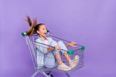 Photo of excited pretty small schoolgirl dressed jeans shirt driving shopping tray empty space isolated violet color backgroundの写真素材