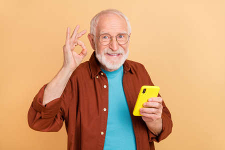 Photo of friendly grandpa use smartphone showing okay symbol recommend product isolated on beige...